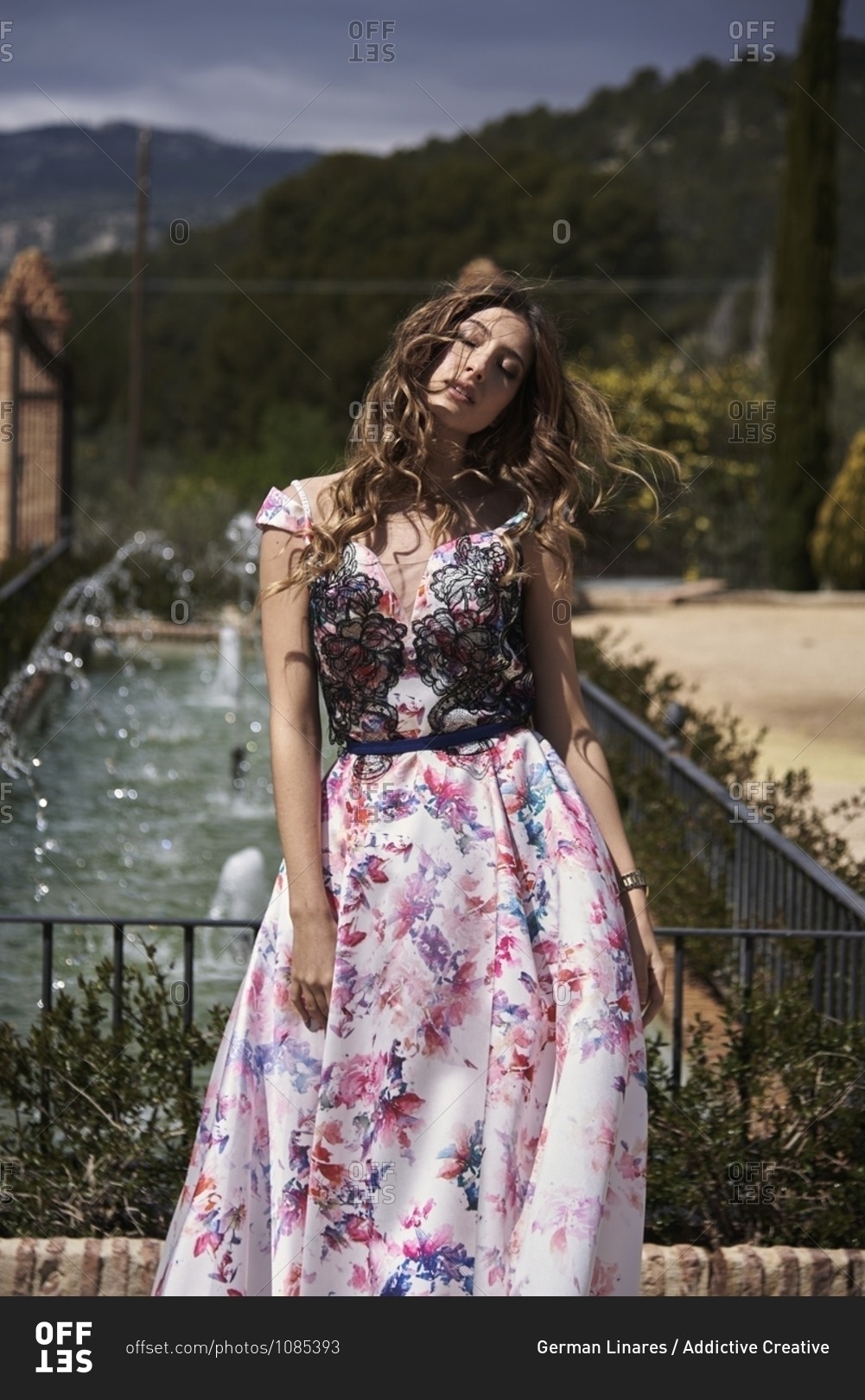 Tender female with wavy hair and in long elegant dress standing in garden near fountain and closed eyes