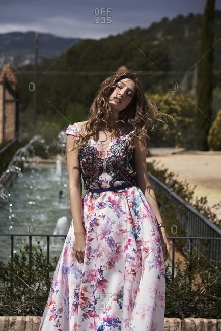 Tender female with wavy hair and in long elegant dress standing in garden near fountain and closed eyes