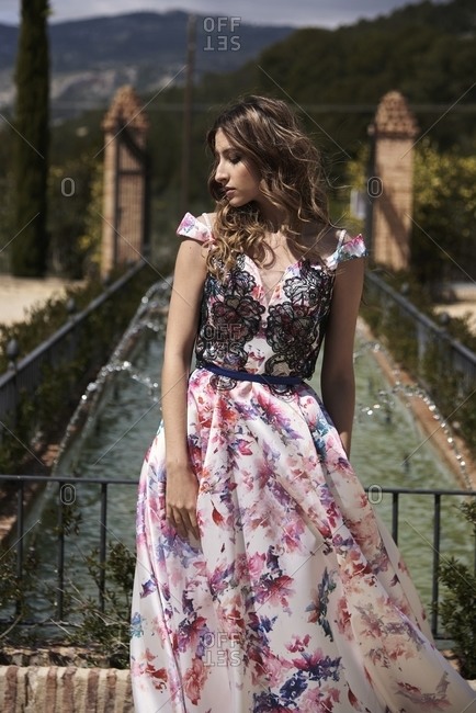 Tender female with wavy hair and in long elegant dress standing in garden near fountain and looking away