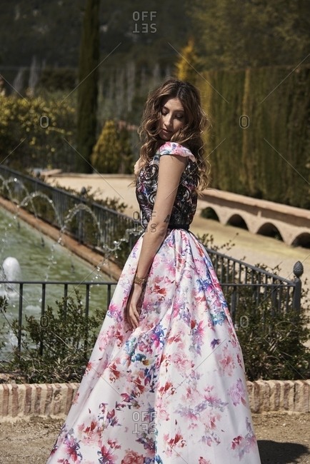 Tender female with wavy hair and in long elegant dress standing in garden near fountain and looking down