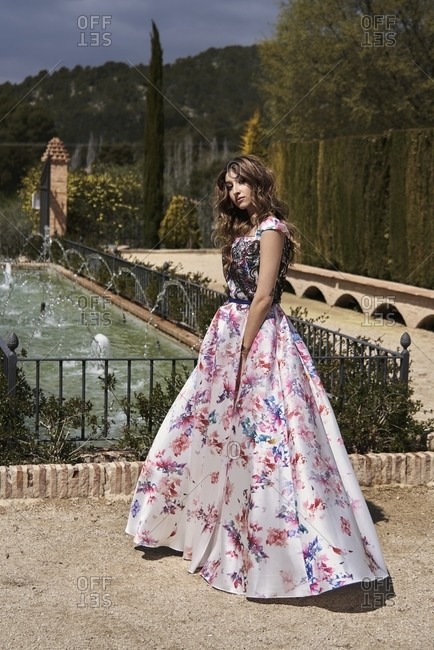 Tender female with wavy hair and in long elegant dress standing in garden near fountain and looking at camera