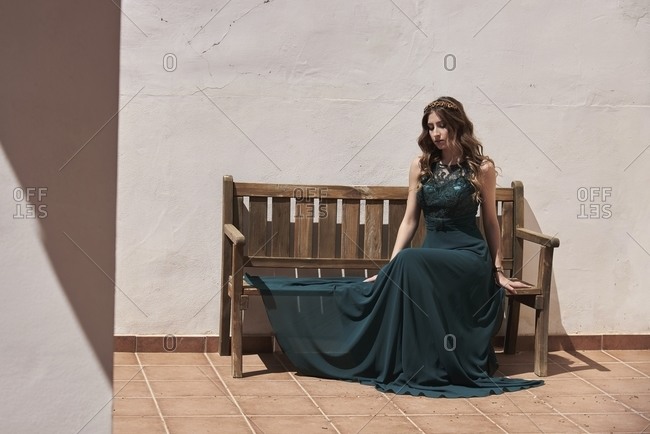 Young peaceful female with long wavy hair and in emerald elegant dress sitting on wooden bench in summer and looking down