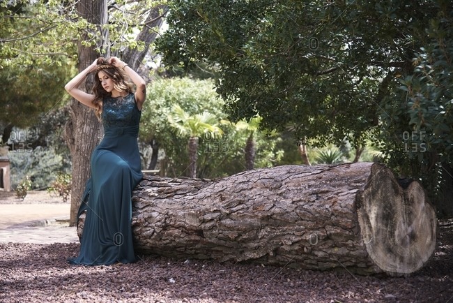 Full body of dreamy female wearing maxi emerald dress sitting on tree trunk in woods and looking down