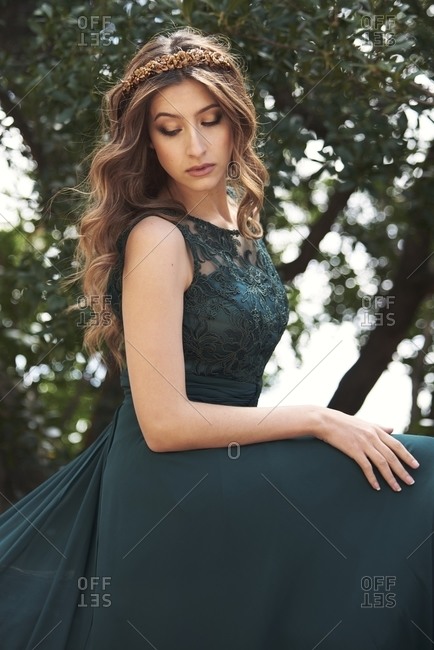 Gentle female in elegant emerald dress and flower hair band standing in street in summer and looking down