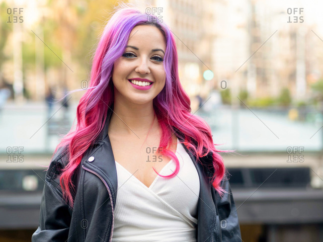 Optimistic confident millennial female with dyed pink hair wearing leather jacket looking at camera and smiling while standing against blurred urban background