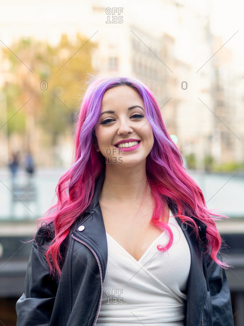Optimistic confident millennial female with dyed pink hair wearing leather jacket looking at camera and smiling while standing against blurred urban background