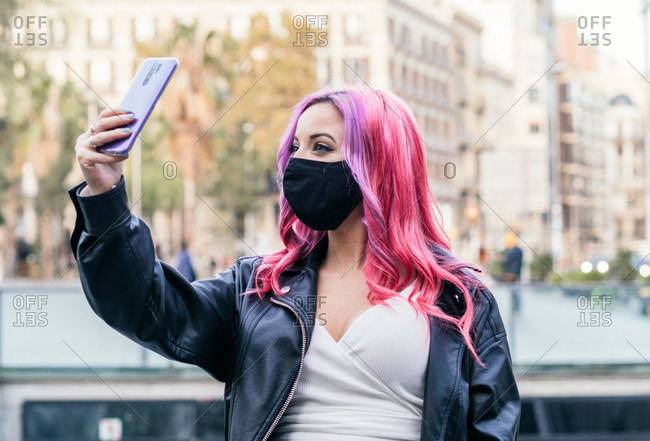 Positive millennial female student with dyed pink hair wearing leather jacket and black protective mask taking selfie on smartphone while standing on city street