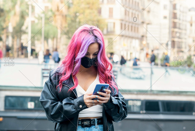 Positive millennial female student with dyed pink hair wearing leather jacket and black protective mask browsing on smartphone while standing on city street