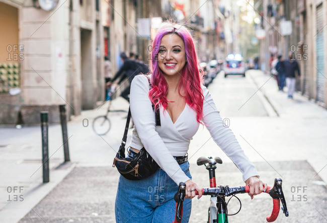 Cheerful young alternative female student with dyed pink hair standing with bicycle on paved urban street
