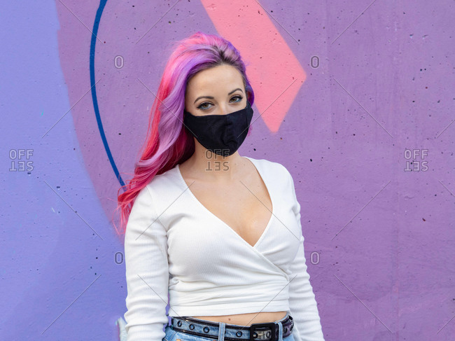 Modern millennial hipster female with dyed long hair wearing black protective mask for coronavirus prevention looking at camera while standing against bright colorful wall