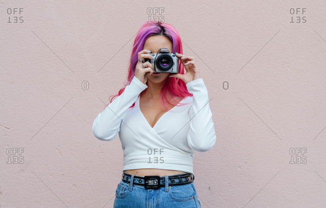 Unrecognizable hipster female with pink hair covering face with camera while shooting photo against beige wall