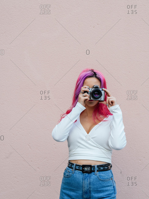 Unrecognizable hipster female with pink hair covering face with camera while shooting photo against beige wall
