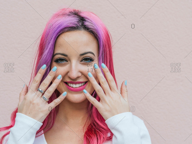 Smiling young female with pink hair and bright makeup showing hands with blue colored nails against beige background