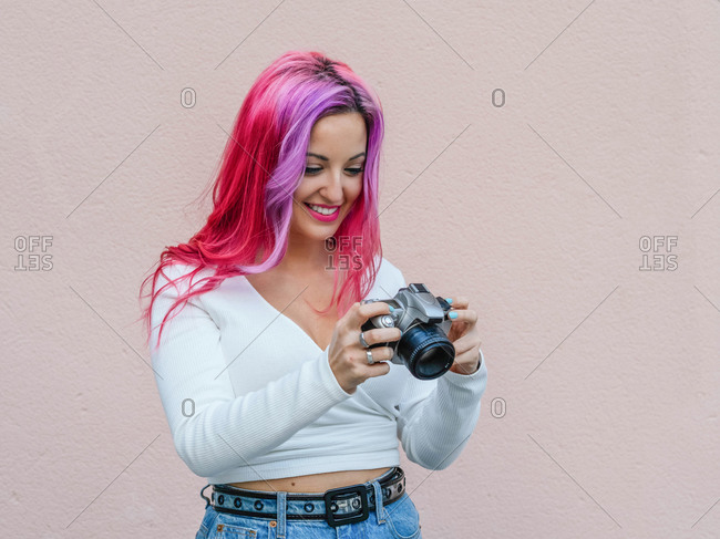 Young hipster female with pink hair looking at camera while shooting photo against beige wall