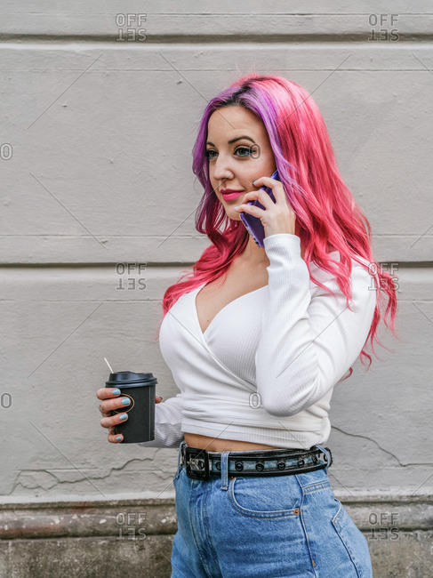 Modern alternative young female with long pink hair holding cup of takeaway coffee and talking on mobile phone while standing near stone wall on street