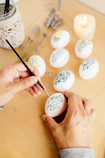 From above of crop unrecognizable person with paintbrush painting flowers on white eggs while preparing for Easter celebration at home