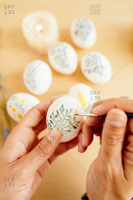 From above of crop unrecognizable person with paintbrush painting flowers on white eggs while preparing for Easter celebration at home