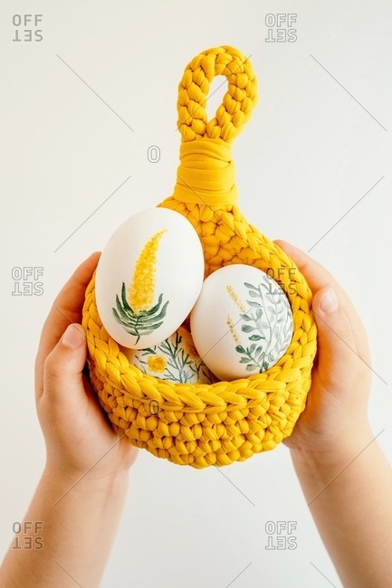 Unrecognizable crop kid with basket of Easter eggs painted with flowers on white isolated background