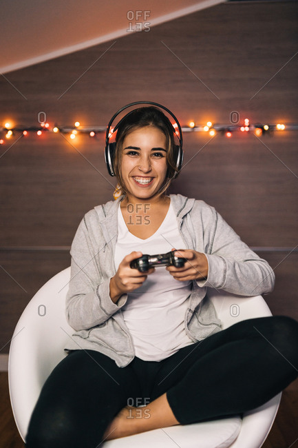 Cheerful female in headphones and with console sitting in armchair and playing video game while entertaining in evening at home