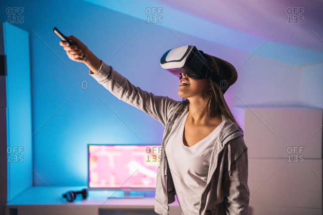Excited female gamer playing video game in virtual reality headset while sitting in room with neon light