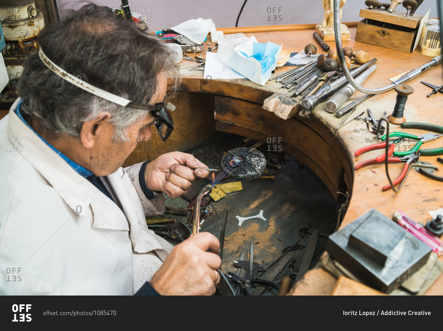 High angle side view of skilled mature male master in glasses using soldering iron while working at workbench with professional instruments in jewelry workshop