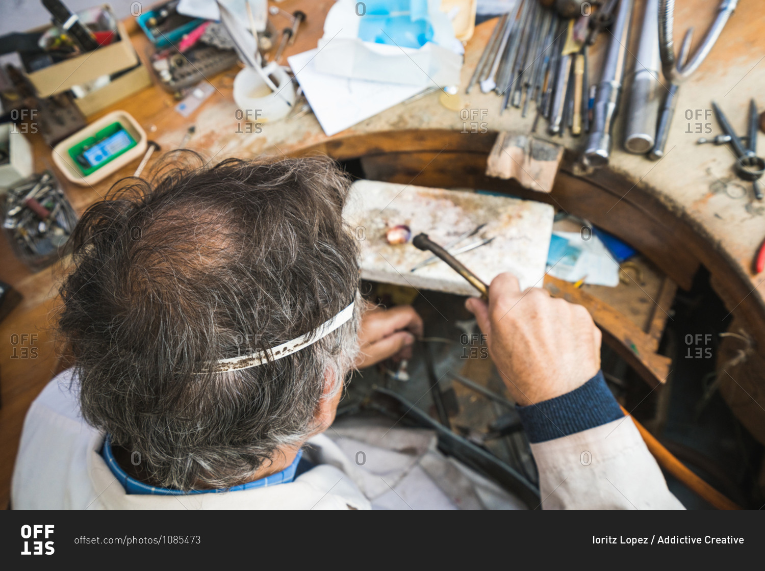 From above of focused skilled mature male master in glasses using hammer and chisel while working with gemstone in jewelry workshop