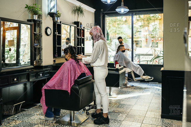 Side view of female hairdresser using curling iron and making trendy hairstyle for client in modern salon during coronavirus pandemic