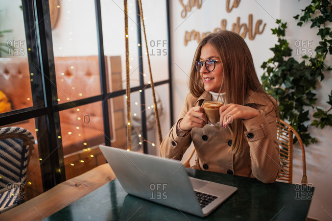 Smiling young female blogger in casual clothes and glasses sitting at table with cup of coffee and typing on laptop keyboard while preparing content for social networks in cozy cafeteria