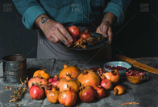 Unrecognizable tattoo hand of person standing at rustic table and putting on ripe fresh pomegranates in pile in dark room