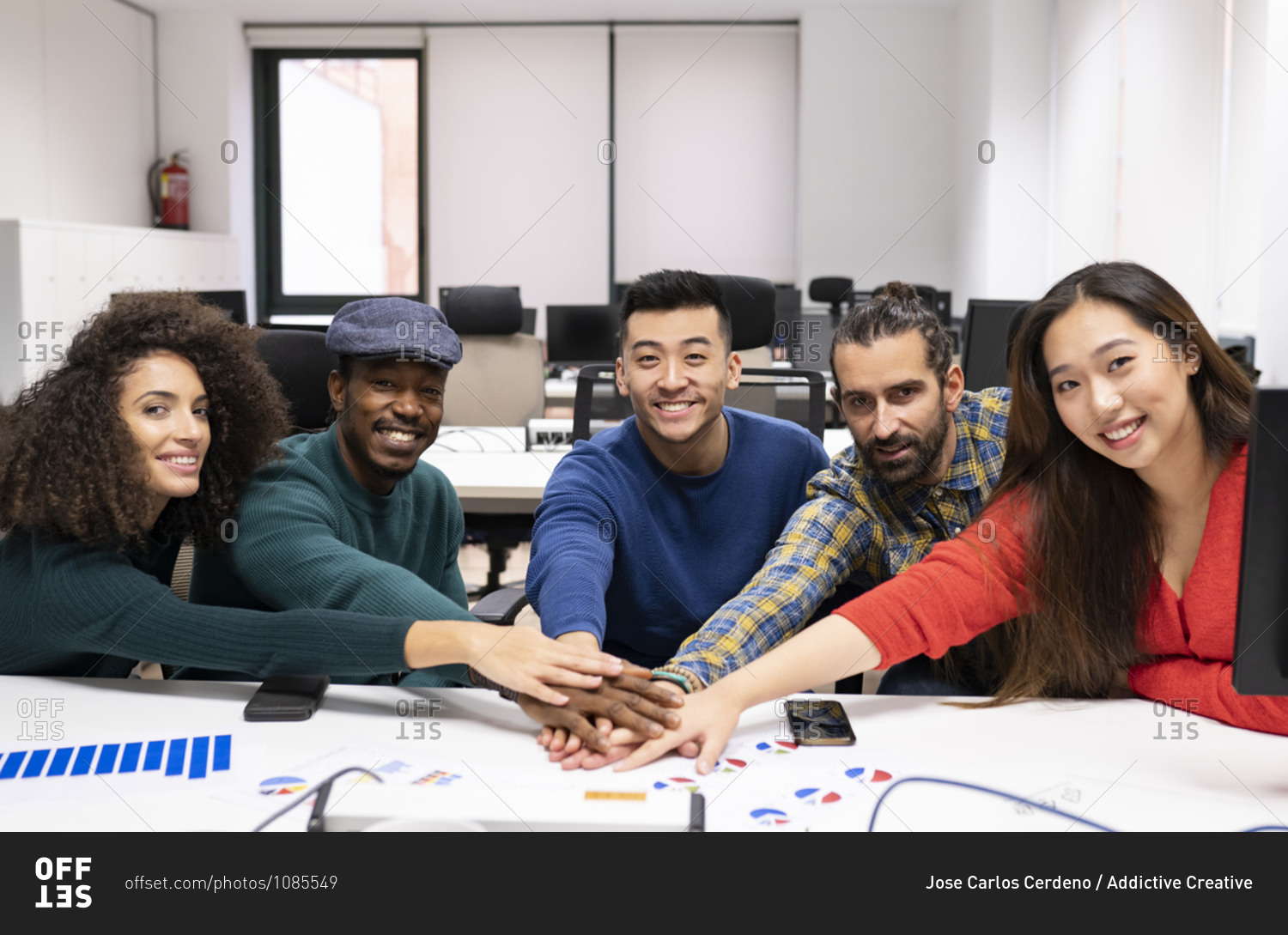 Young multiracial successful startup team gathering around table with diagrams and stacking hands together while discussing business strategy in modern office
