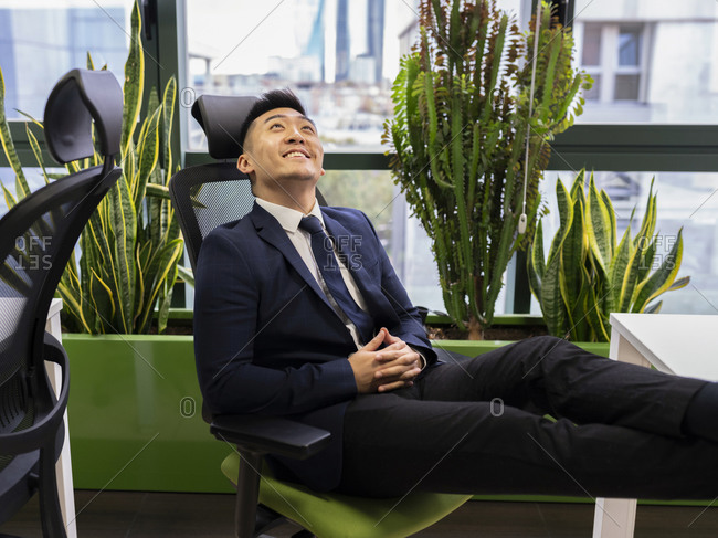 Side view of cheerful young Asian businessman in formal suit looking up and smiling dreamfully while thinking about successful business strategy in modern workspace