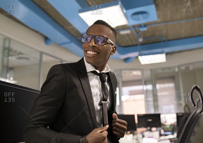 Low angle of positive young African American businessman dressed in elegant black suit with tie and eyeglasses working in contemporary workspace
