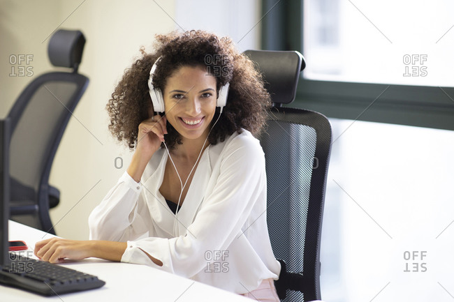 Young Hispanic female employee in white blouse and headphones smiling friendly at camera while working at table with computer in modern workplace