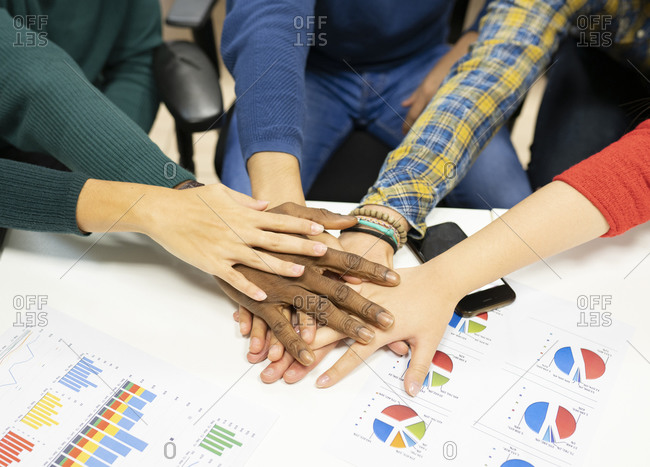 From above of crop anonymous multiracial colleagues in casual clothes gathering around table with charts and stacking hands together while working on business development plan in workplace