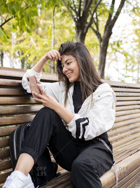 Cheerful young Latin female in sporty outfit sitting on bench and browsing mobile phone while resting in park