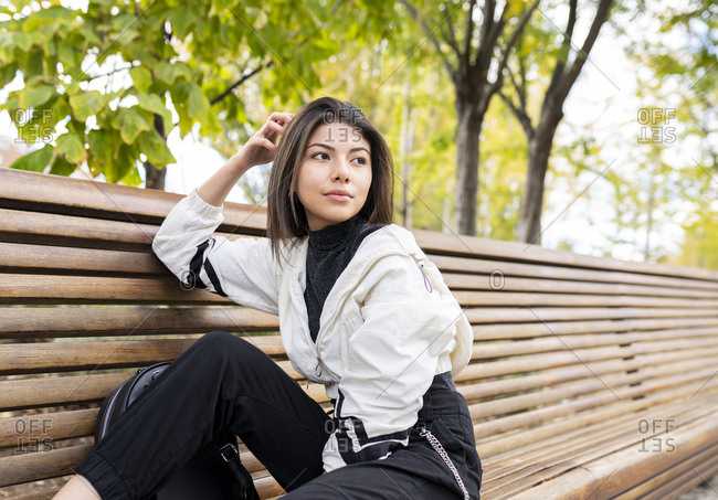 Side view of tranquil dreamy young Latin female in stylish spotty outfit sitting on wooden bench and looking away thoughtfully while resting in park