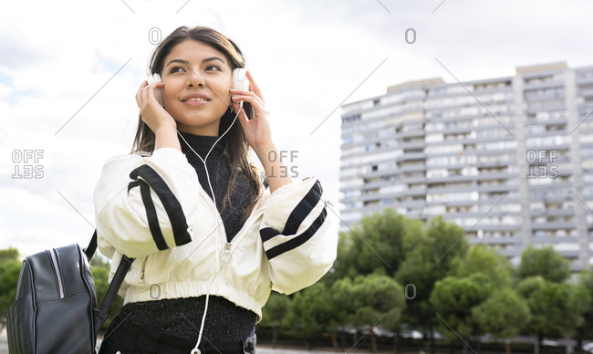 From below of positive young ethnic female in casual outfit listening to music with headphones and browsing mobile phone while chilling in park
