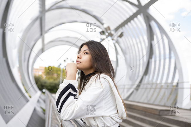 Side view of lonely contemplative young Latin female in trendy casual outfit standing on modern footbridge and looking away thoughtfully