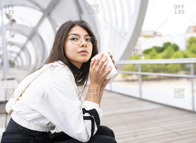 Side view of young Latin female student in casual outfit and glasses drinking takeaway coffee and looking away thoughtfully while chilling on footbridge over river in city