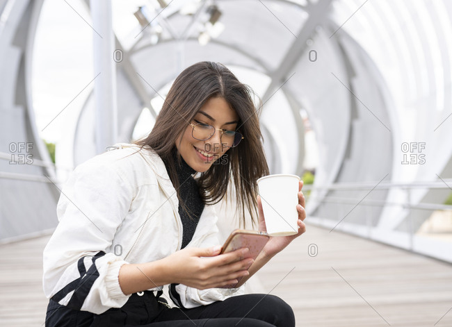 Side view of positive young ethnic female student in casual outfit and eyeglasses browsing mobile phone while resting with cup of takeaway coffee on enclosed pedestrian bridge