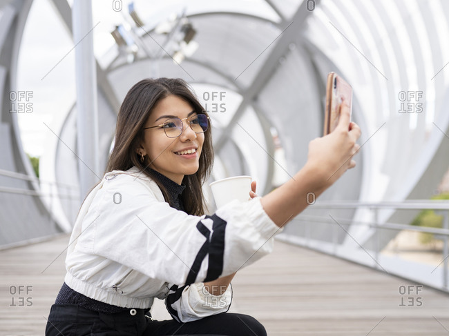Side view of positive young ethnic female student in casual outfit and eyeglasses taking selfie on mobile phone while resting with cup of takeaway coffee on enclosed pedestrian bridge