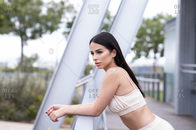 Side view of young slim brunette female in stylish sportswear leaning on railing and looking away while resting after outdoor workout