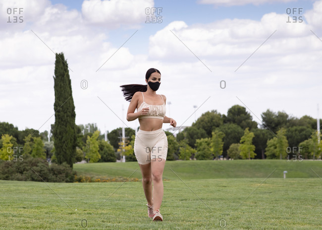 Full body of young fit female in trendy activewear and mask running on green grassy meadow towards during fitness training in park