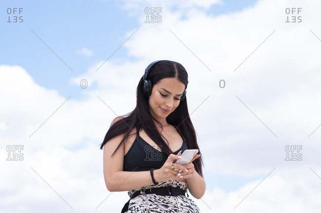 Positive young fit female in sports outfit listening to music through wireless headphones and browsing mobile phone while standing against blue cloudy sky and resting after outdoor workout