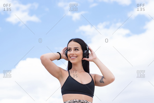 Positive young fit female in sports outfit listening to music through wireless headphones while standing against blue cloudy sky and resting after outdoor workout
