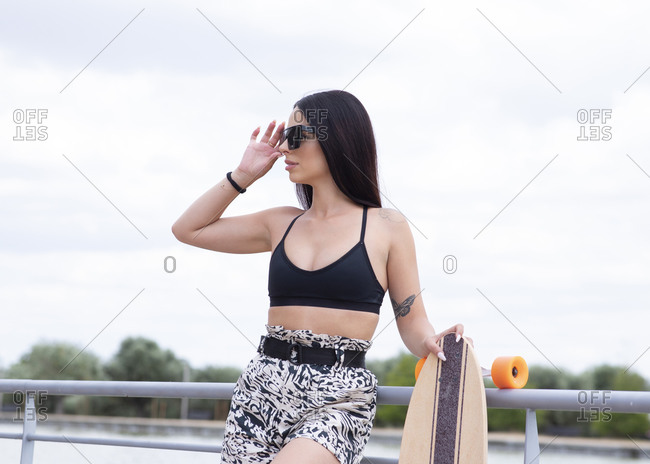 Confident young slim brunette in stylish activewear and trendy sunglasses holding longboard and looking away while standing near railing on embankment