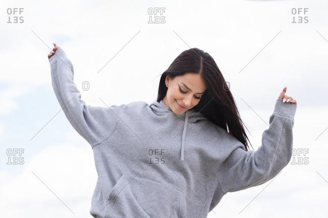 Low angle of happy young female in gray hoodie having fun and jumping with arms raised against cloudy sky