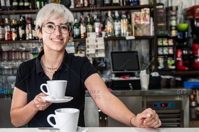 Cheerful female bartender standing at counter with cup of freshly prepared coffee and looking at camera