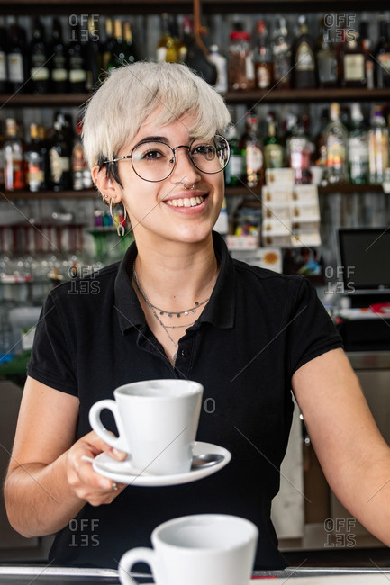 Cheerful female bartender standing at counter with cup of freshly prepared coffee and looking at camera