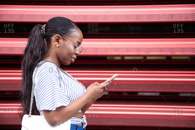 Side view of positive young black female student in casual clothes and eyeglasses browsing mobile phone while standing near red fence on street